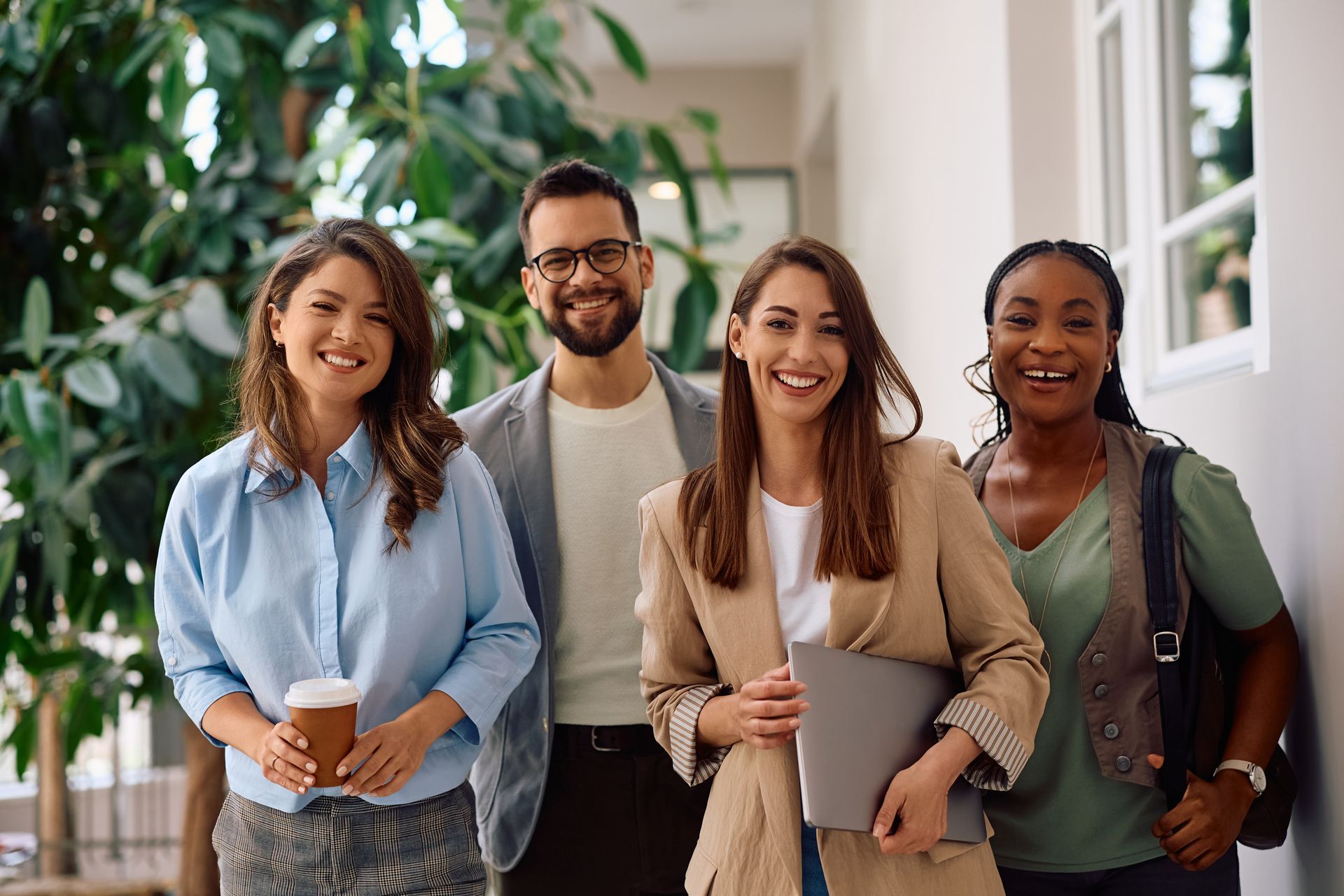 Four diverse office workers smiling at the camera. Hallway setting with plants and natural light.