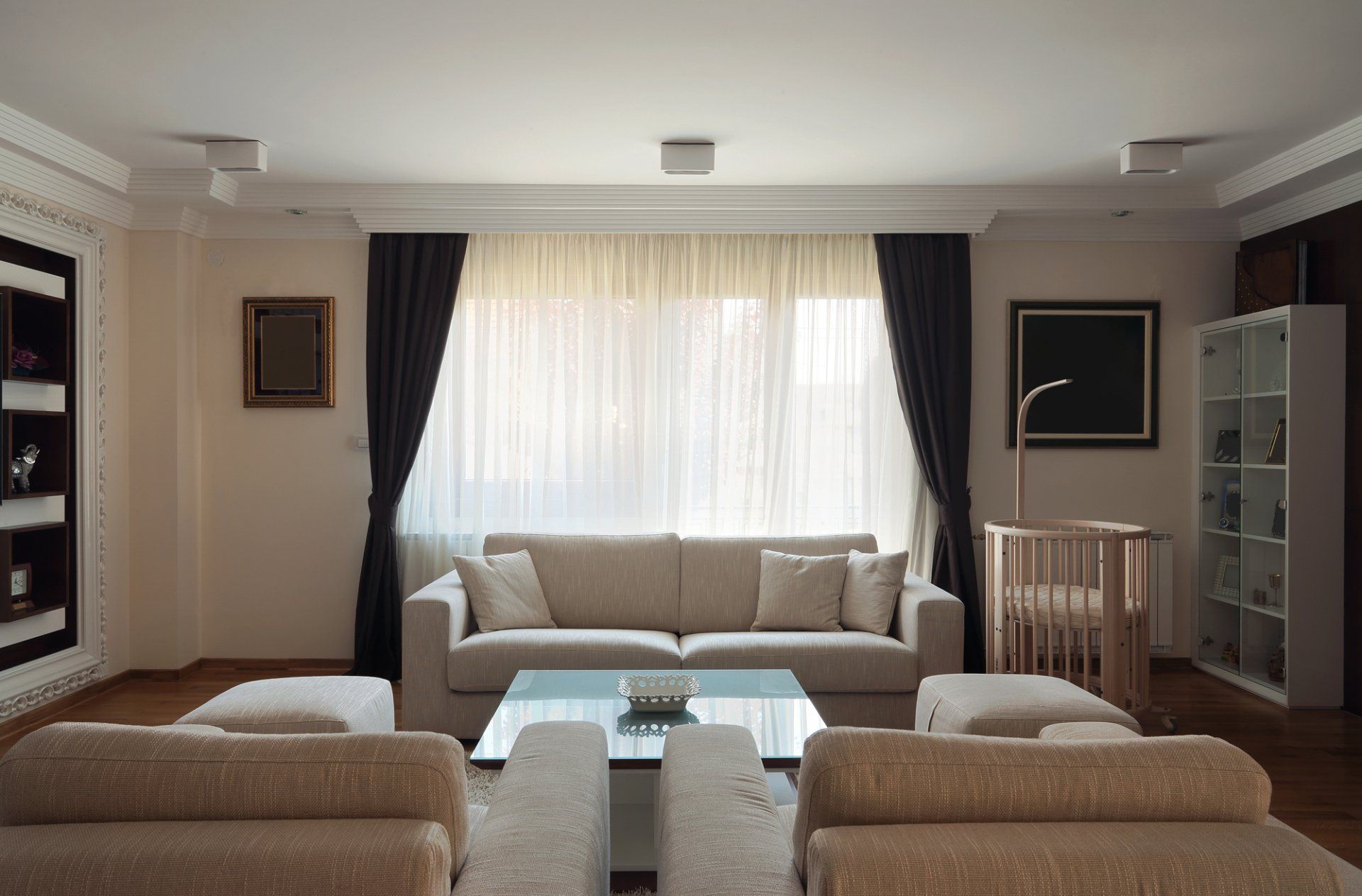 Neutral toned loungeroom with couch and chairs around coffee table. Dark formal curtains pulled tied back to expose white sheer curtains across window.
