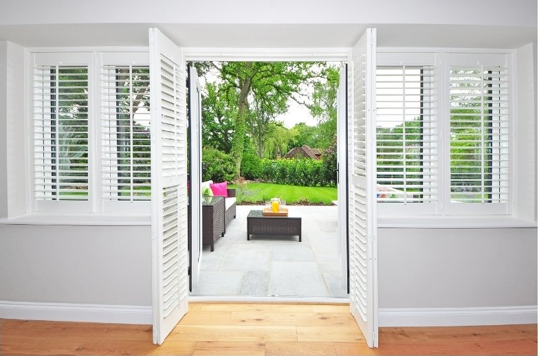 View out open french doors with open plantation shutters, looking out over patio and green garden.