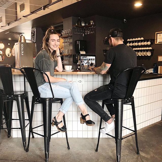 Debbie and Gabriel Mayes sit at the counter of a coffee shop while Debbie smiles at the camera and Gabrielle, on his laptop, learns how email lists can increase profits in your business