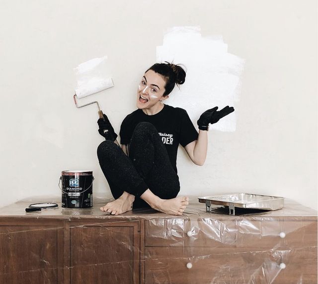 Debbie Mayes sit on top of a wooden chest of draws as she poses with a paint roller as she gets ready to paint the wall white during The Mayes Team's renovation on a budget.