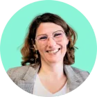 Femme avec des lunettes souriante, portant un blazer et une chemise blanche, sur un fond bleu sarcelle.