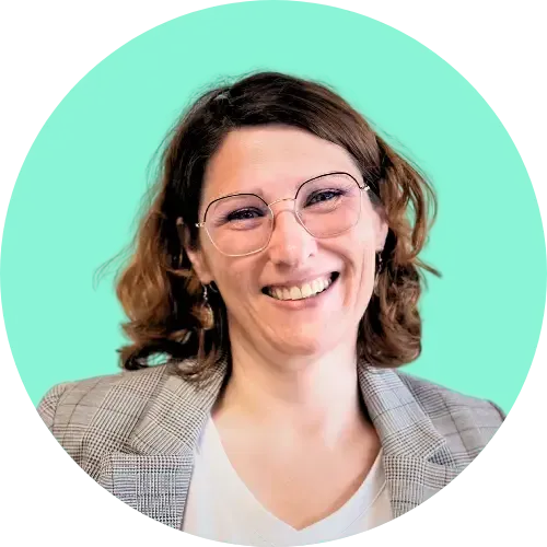 Femme avec des lunettes souriante, portant un blazer et une chemise blanche, sur un fond bleu sarcelle.