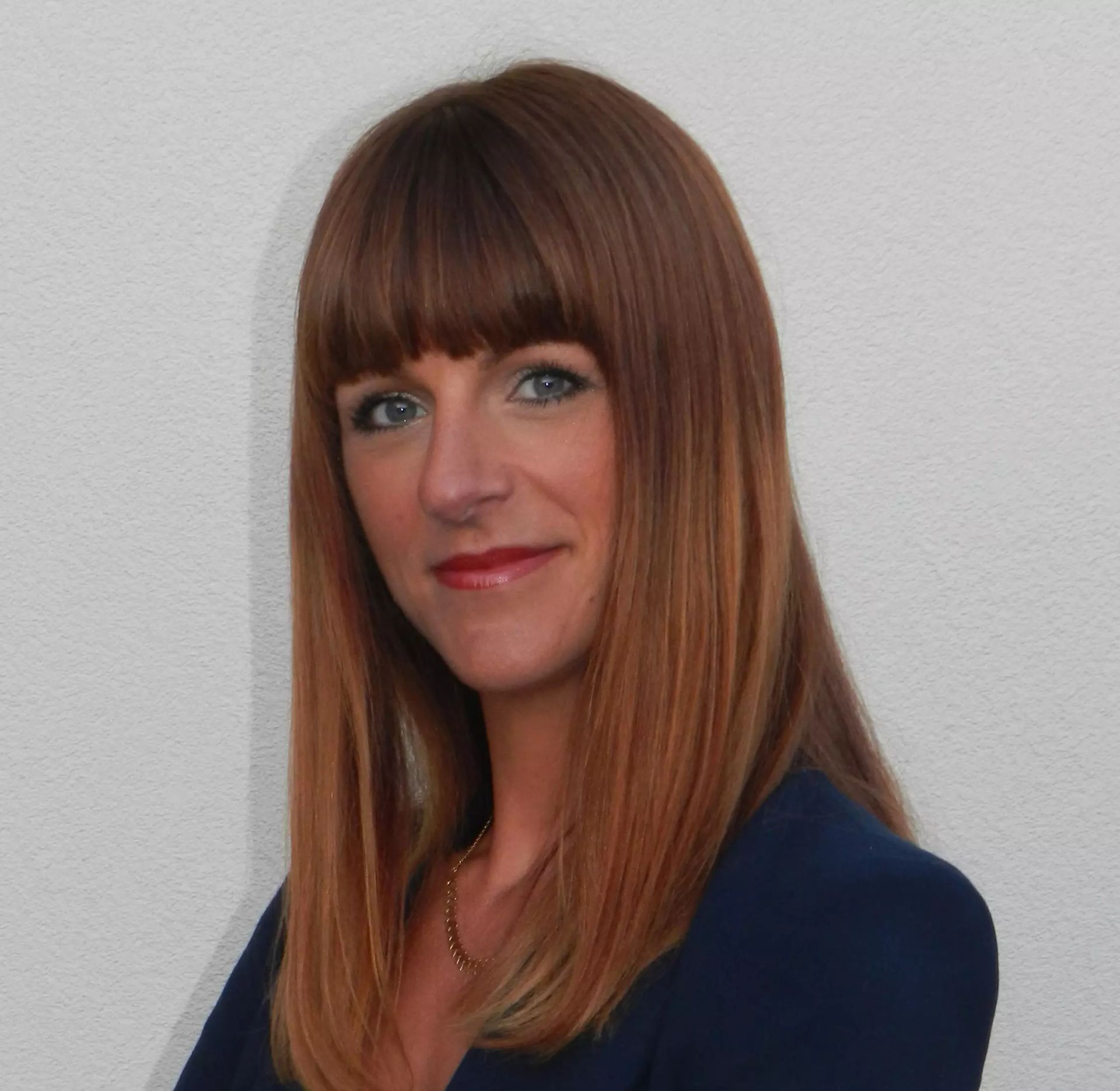 Une femme aux cheveux auburn et à la frange, portant un blazer bleu foncé, sourit.