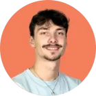Jeune homme souriant avec une moustache, une chemise bleu clair et un collier, sur fond orange.