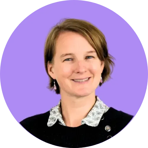 Une femme aux cheveux bruns courts sourit, vêtue d'un pull noir avec un col blanc à motifs sur fond violet.