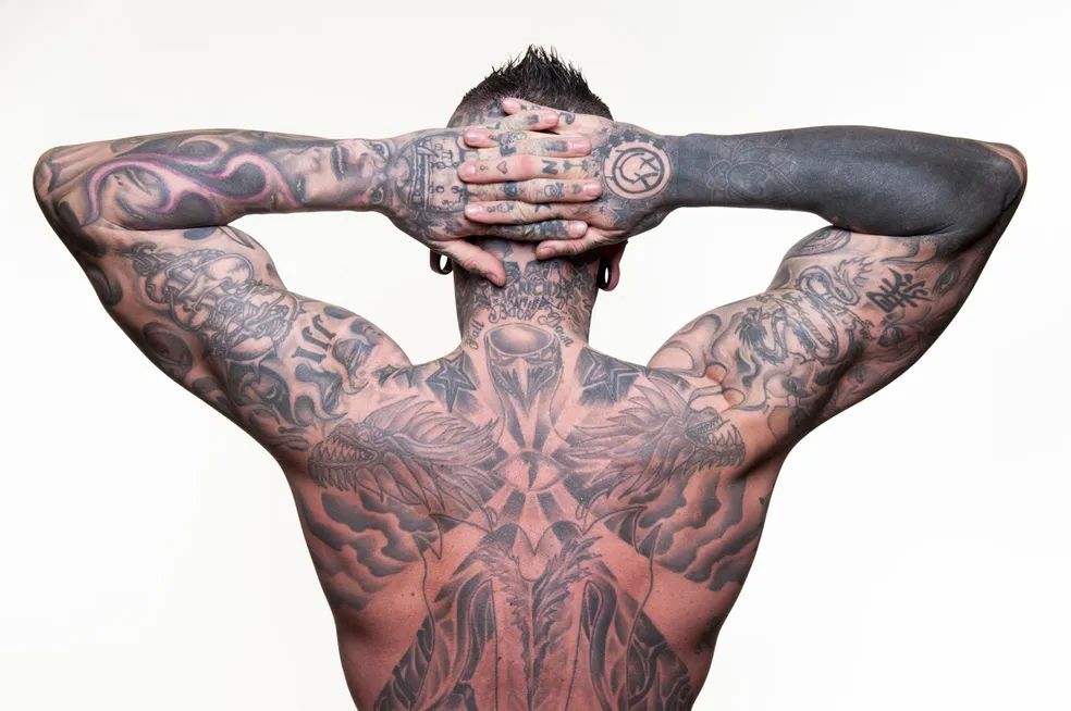 A man with a lot of tattoos on his back is standing with his hands behind his head.