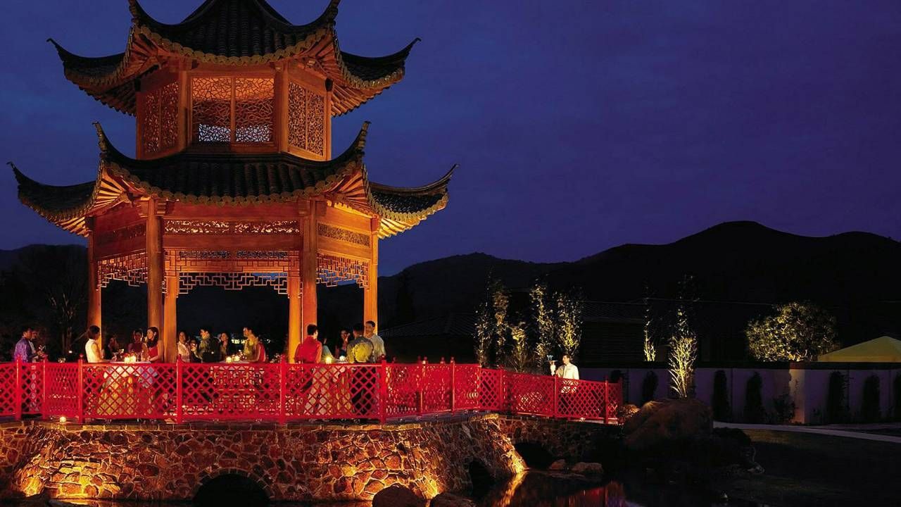 Lit, ornate Chinese pagoda at night, over water, with figures on a red bridge.