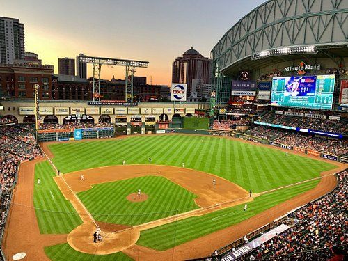 Baseball stadium at sunset, green field, players on the field, fans in stands, city skyline in background.