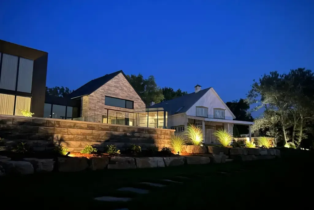 Illuminated homes with stone facades at dusk, surrounded by landscaping and trees.