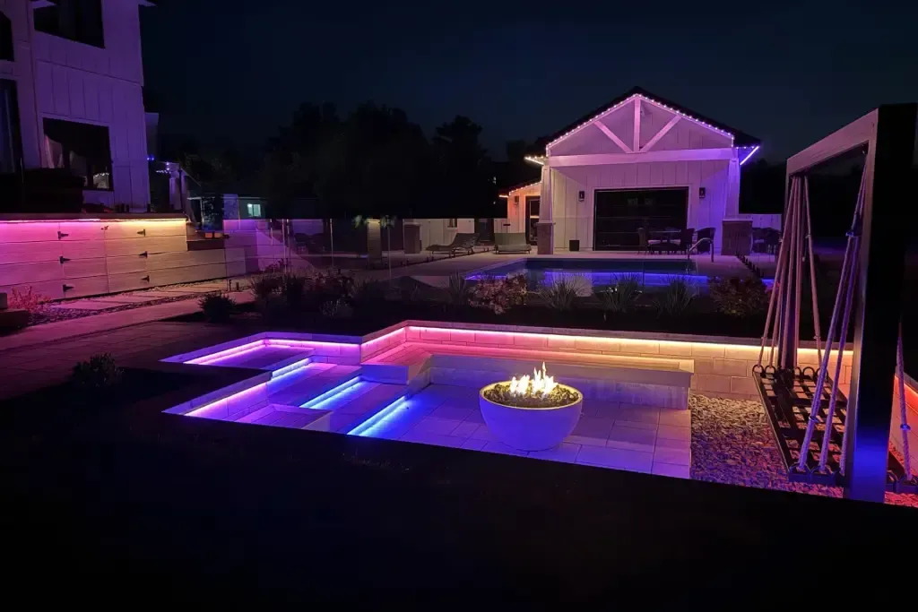 Pool at night lit with colorful lights; house and fire pit in the background.