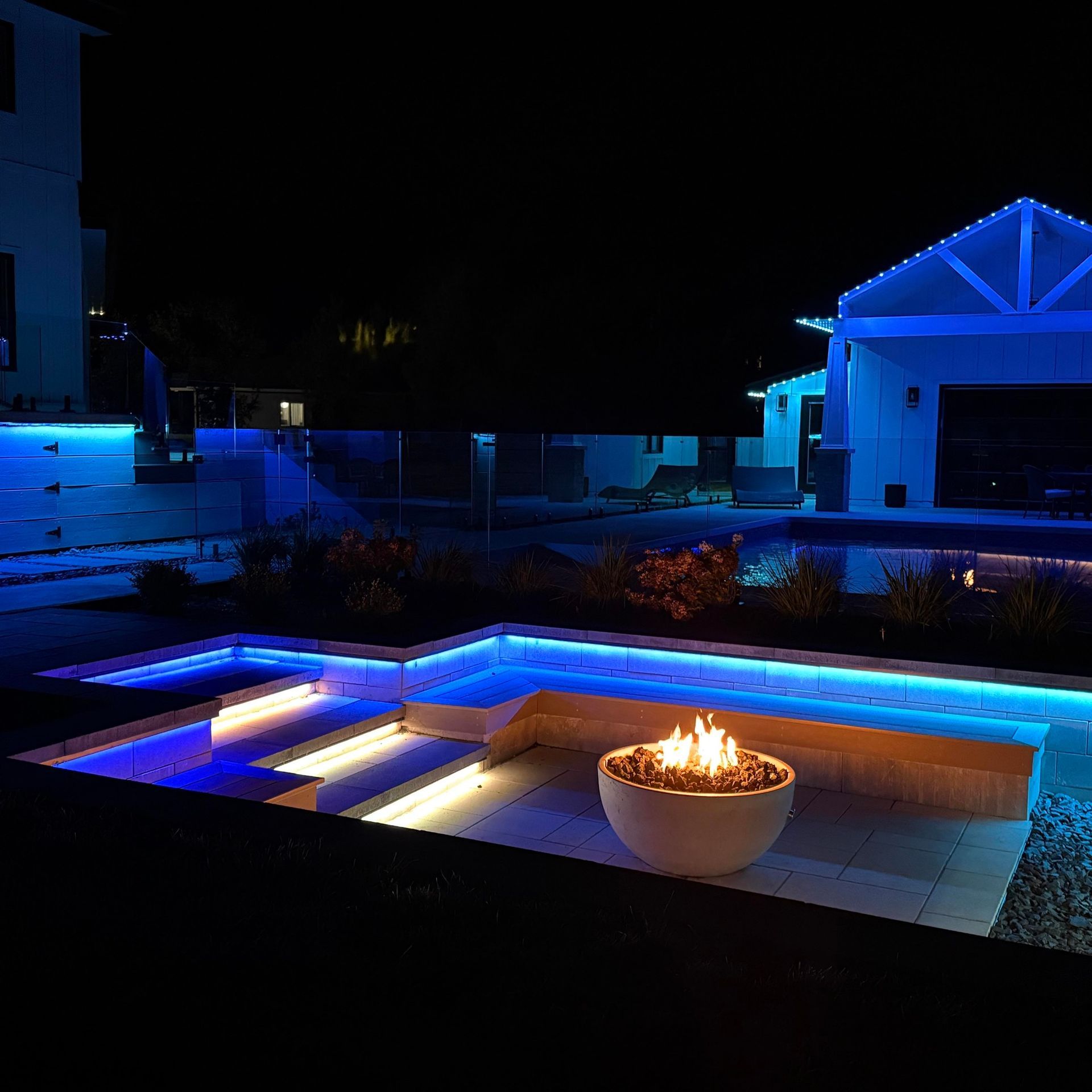 Night view of a modern backyard with blue and white LED lights illuminating a pool, steps, and a fire bowl.