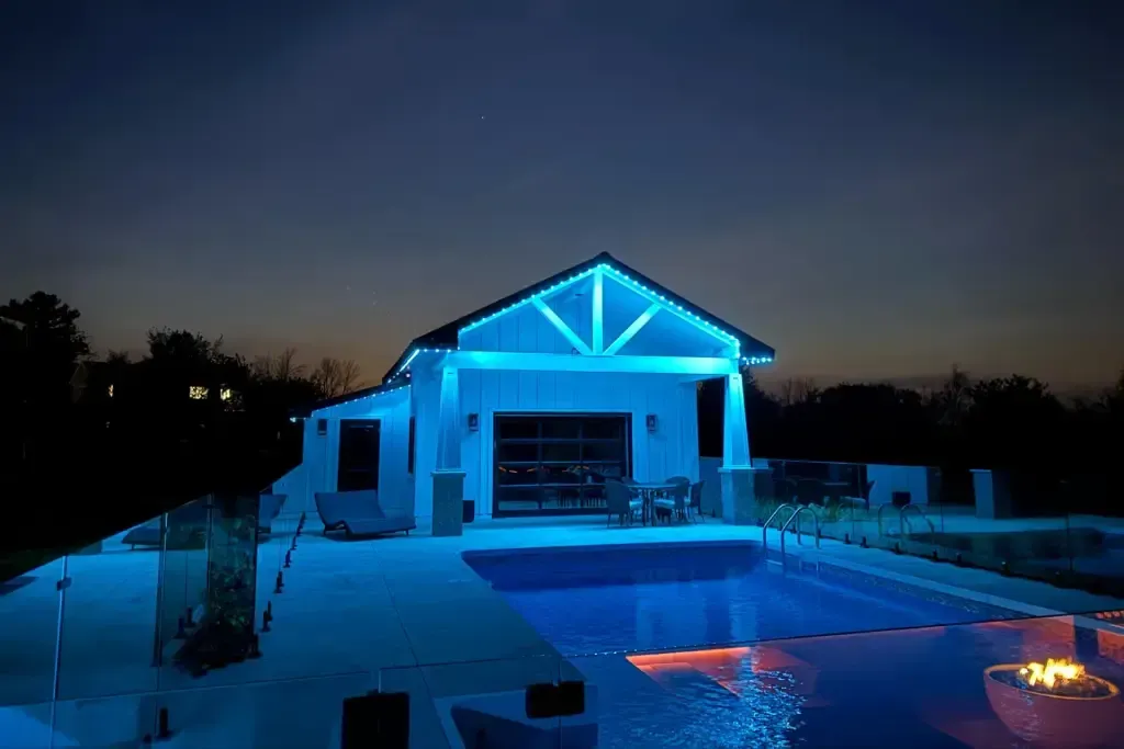 Pool house with blue exterior lighting, a pool, and a fire pit at dusk.