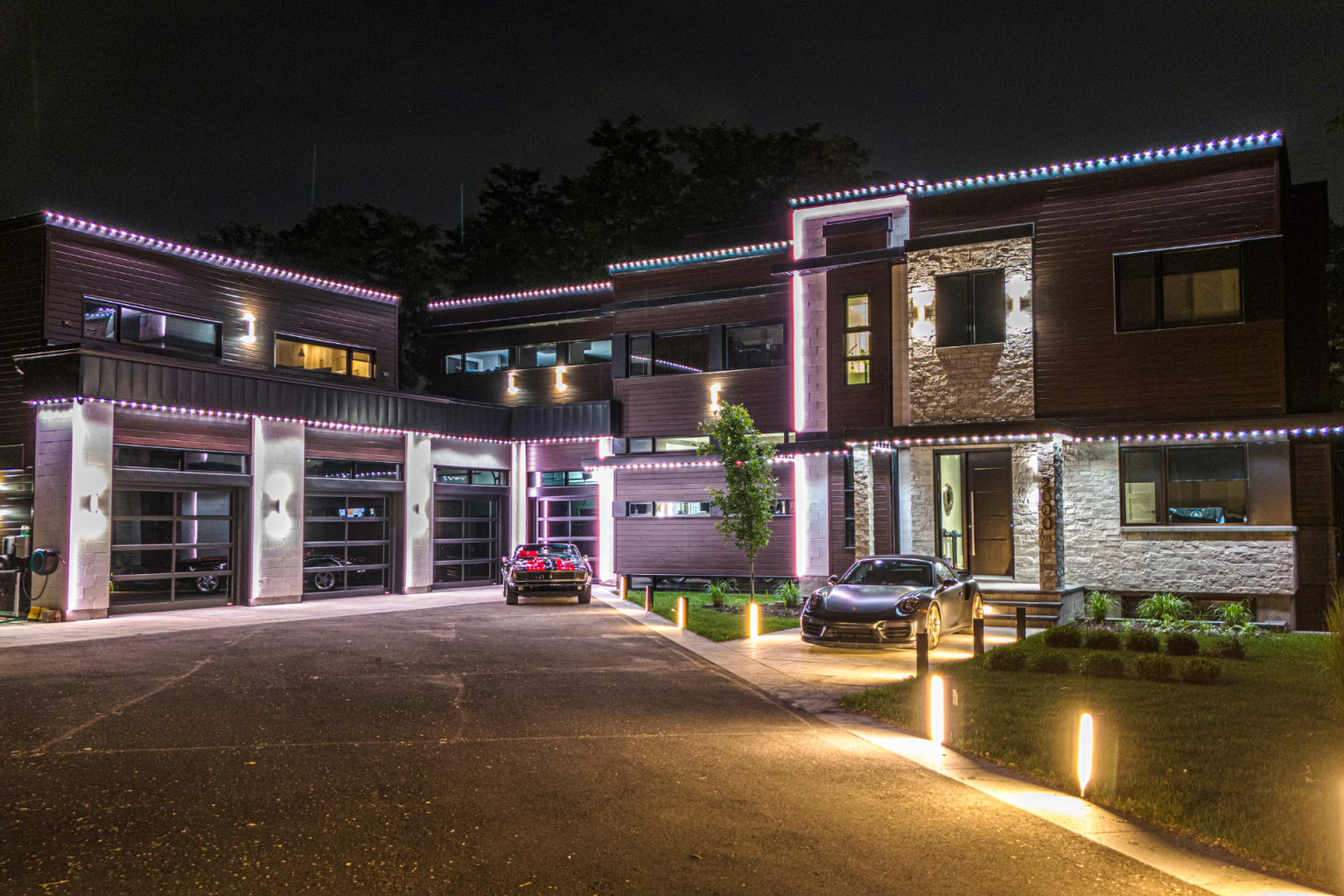 Modern multi-story house lit at night with cars parked in the driveway. Decorative lights are featured.
