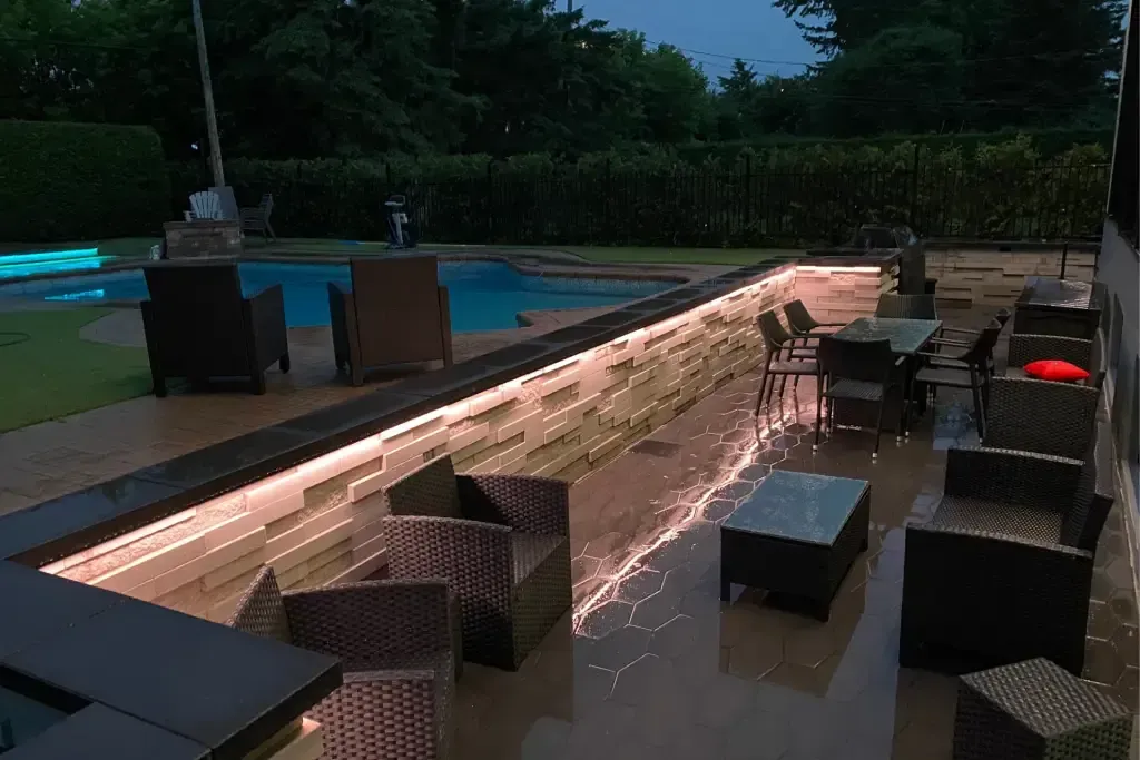 Patio with illuminated ledge, seating, and pool in the background at night.
