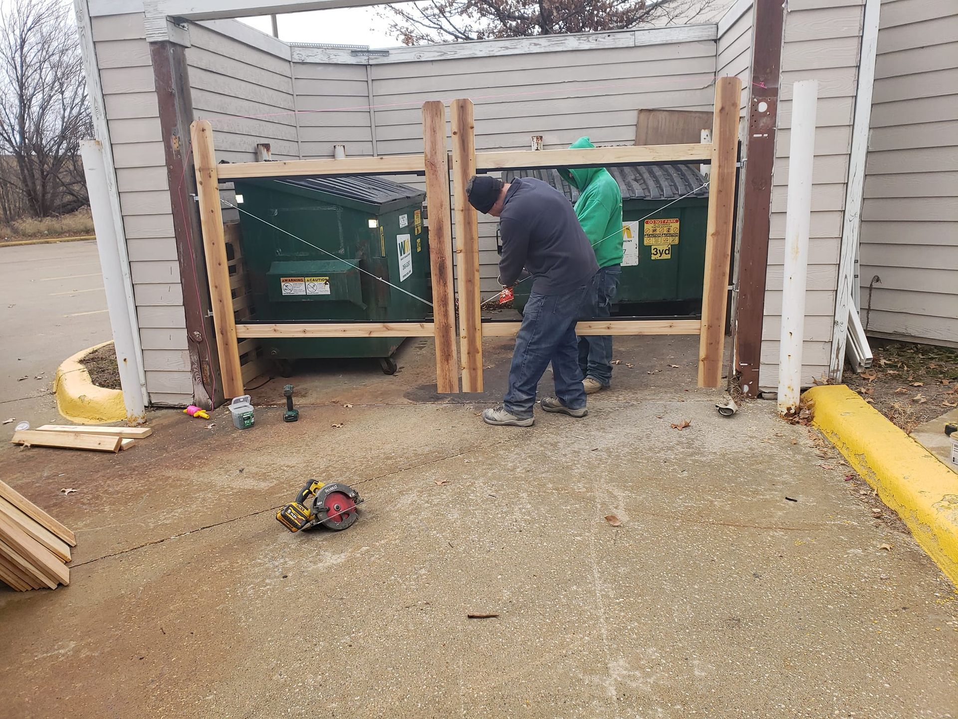 A man is working on a wooden fence in front of a dumpster.