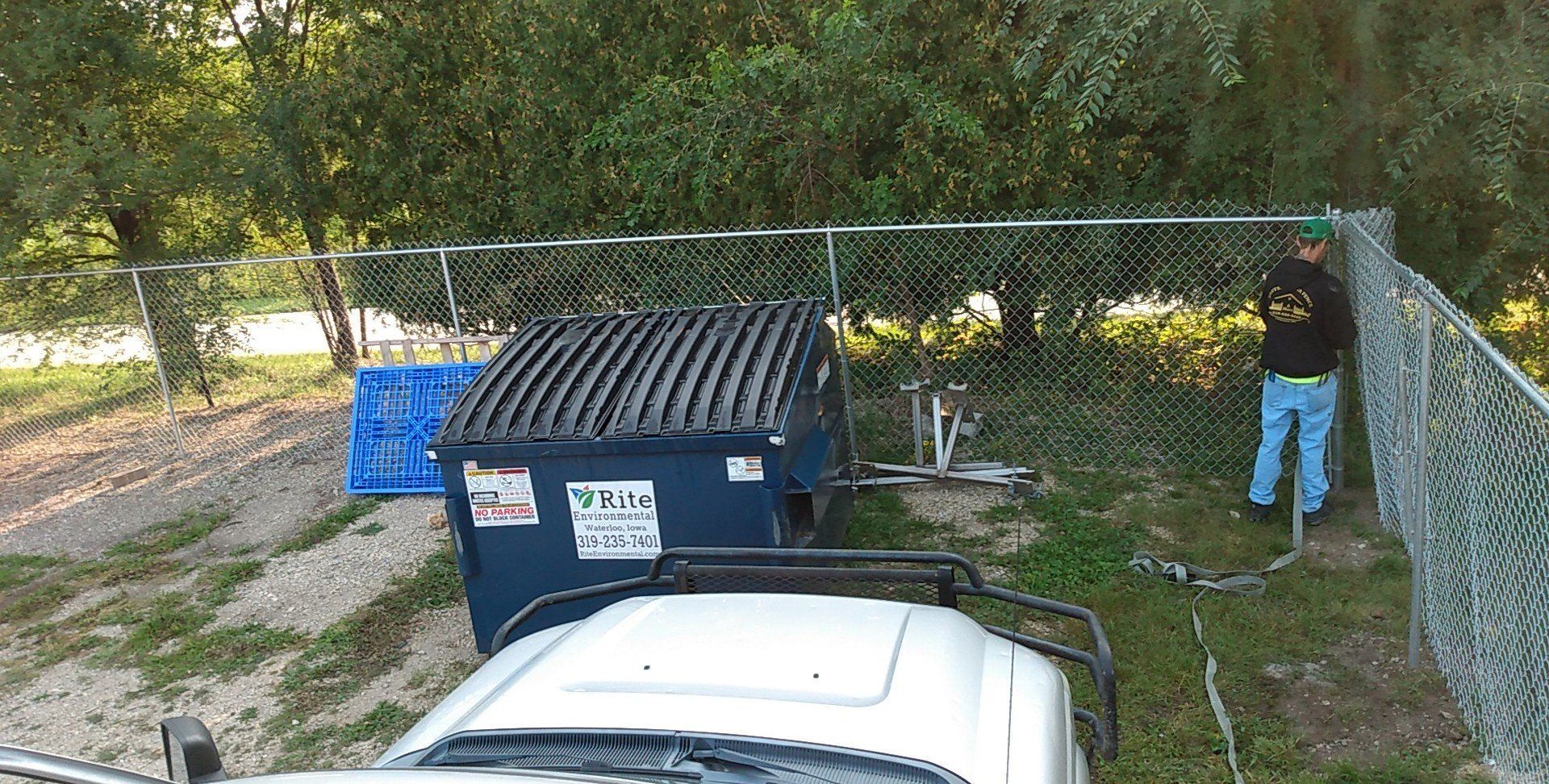 A man is standing next to a fence next to a dumpster.