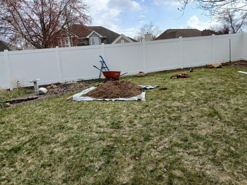 A backyard with a white fence and a wheelbarrow filled with dirt.