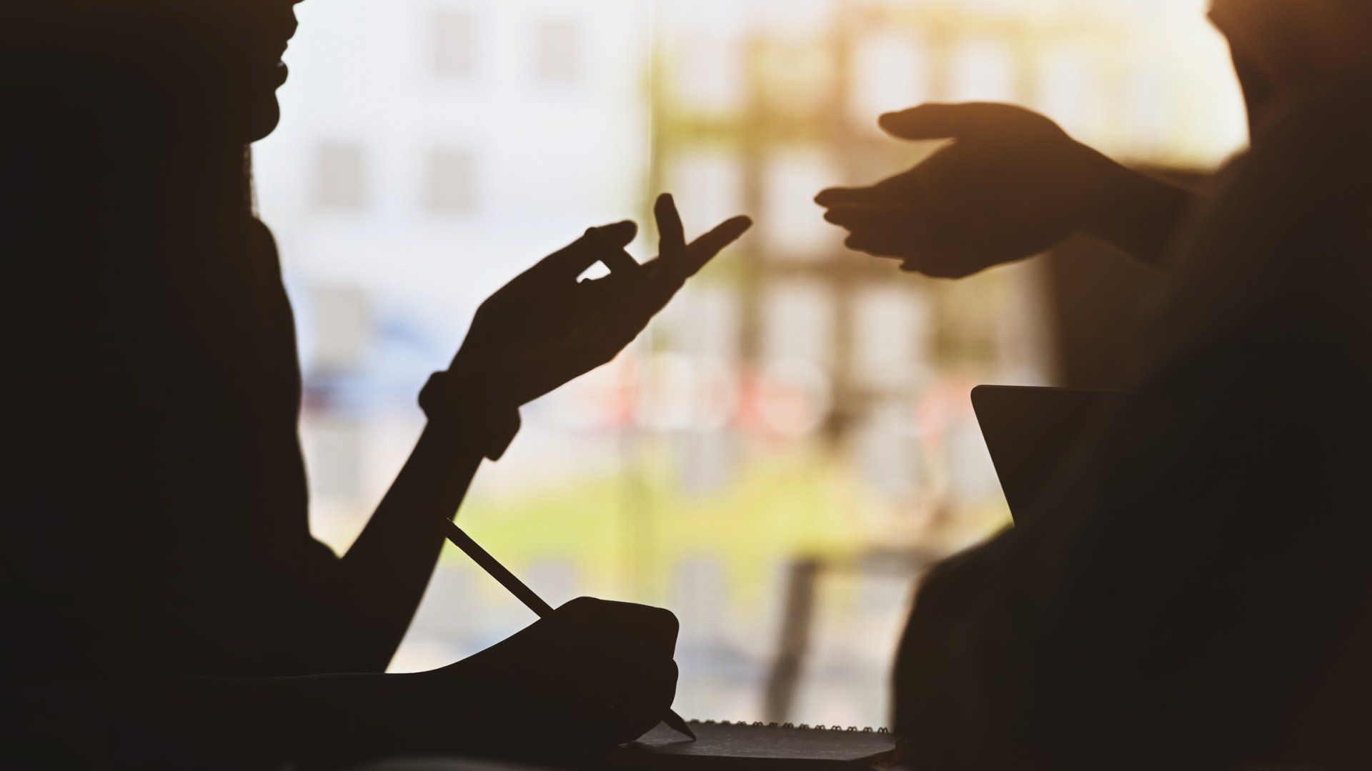 A silhouette of two people talking to each other in front of a window.