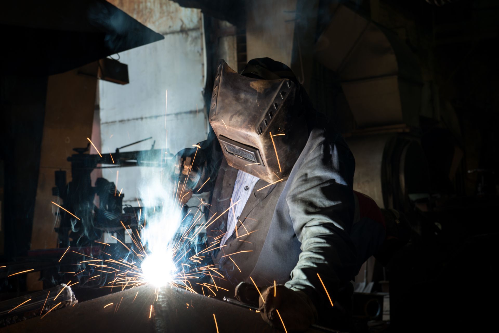 Welder wearing a protective mask, welding with bright sparks in a dimly lit workshop.