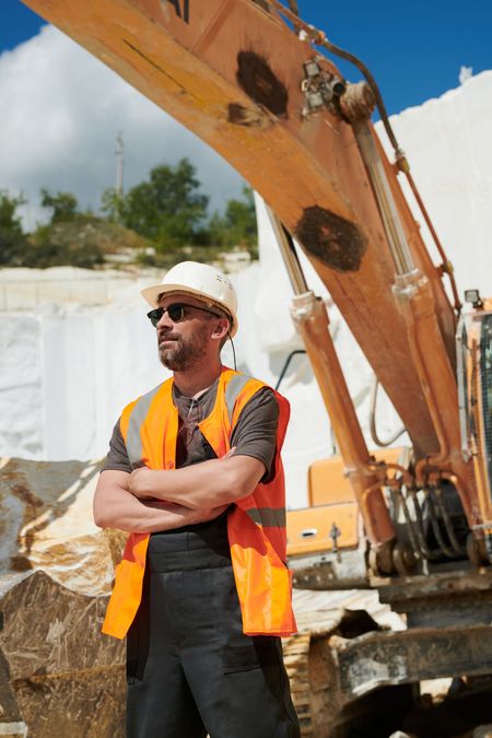 Construction worker in safety gear stands with arms crossed near excavator.