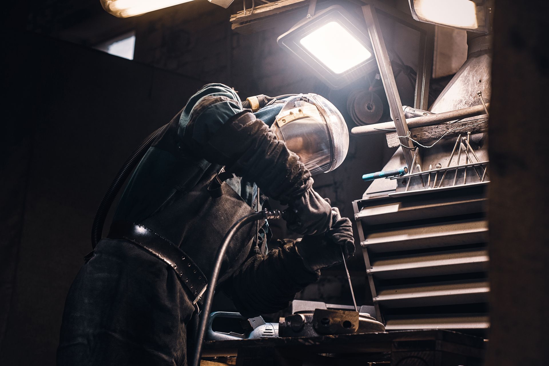 A worker in protective gear welds metal in a dim industrial setting, lit by overhead lamps.