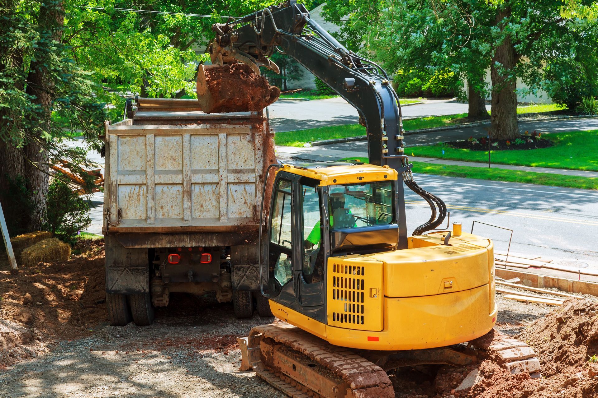 Yellow excavator loading dirt into a dump truck on a street, trees in the background.
