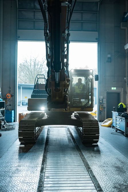 Excavator inside a garage, facing the viewer; bright light shines from the open doorway.