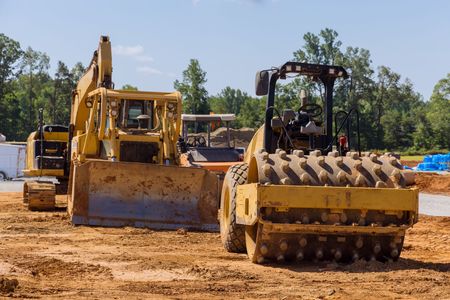 Yellow construction equipment, including a bulldozer and a roller, on a dirt site under a blue sky.