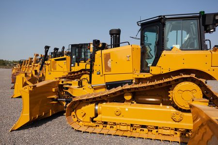 Yellow bulldozers lined up in a row on a gravel lot under a clear sky.