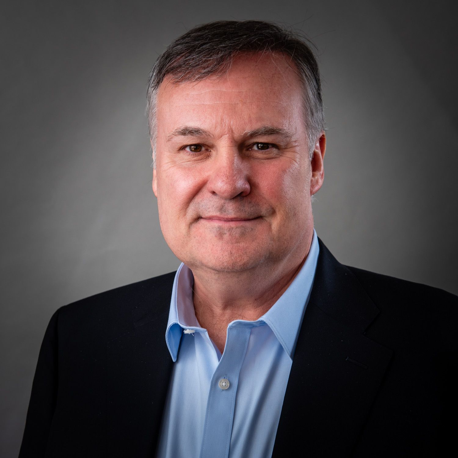 Man in a black suit jacket and light blue shirt smiles at the camera against a gray background.