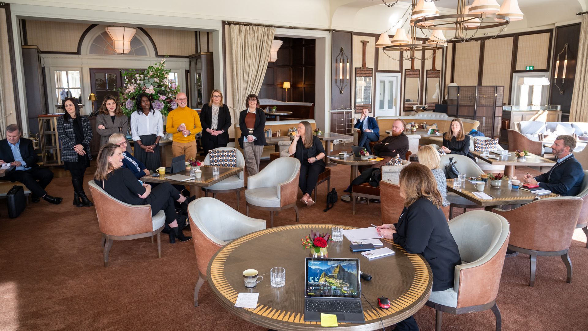 Group of people in a meeting at a cafe, some seated at round tables, others standing.