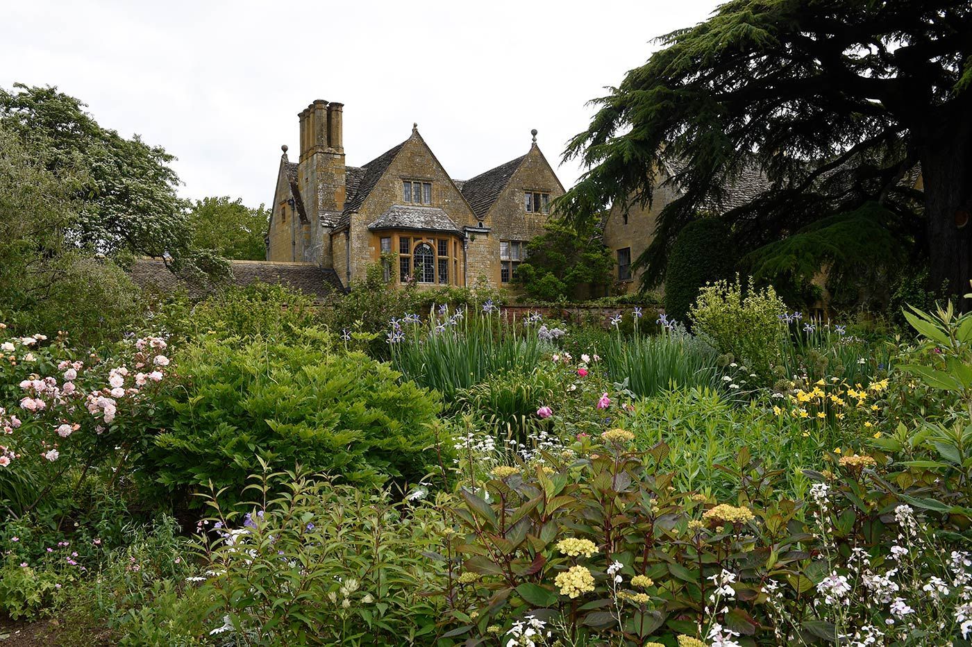 Stone house with a garden foreground. Green foliage, flowering plants, and cloudy sky.