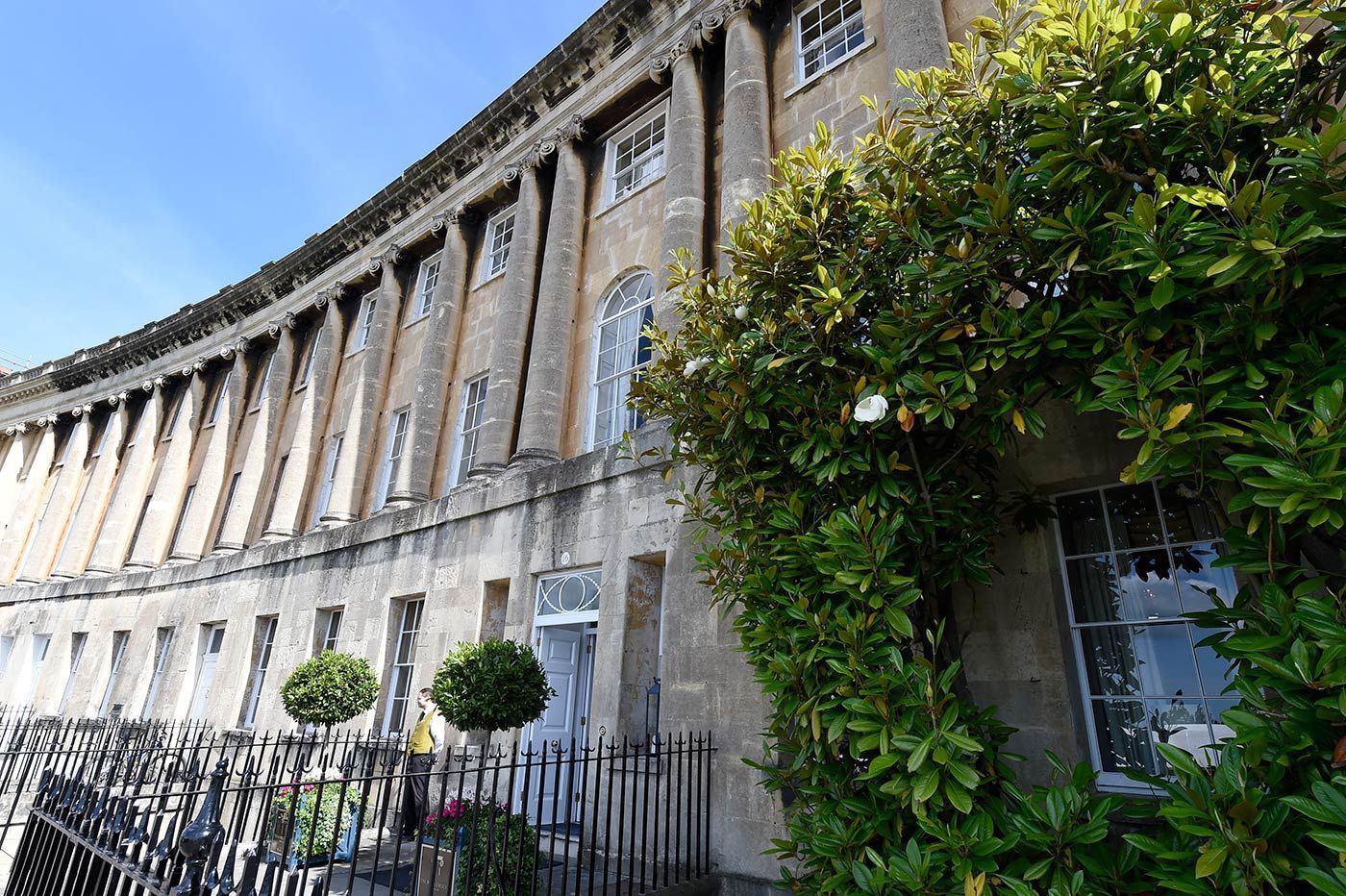 Curved row of pale stone buildings with arched windows, partially obscured by green foliage, under a blue sky.