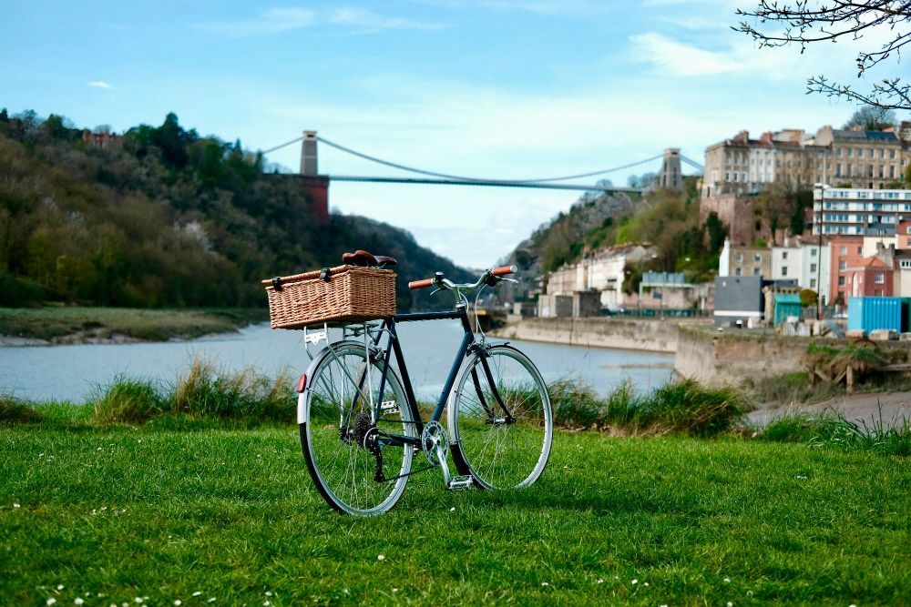 Bicycle with basket on grassy bank, Clifton Suspension Bridge in background, river, and colorful buildings.
