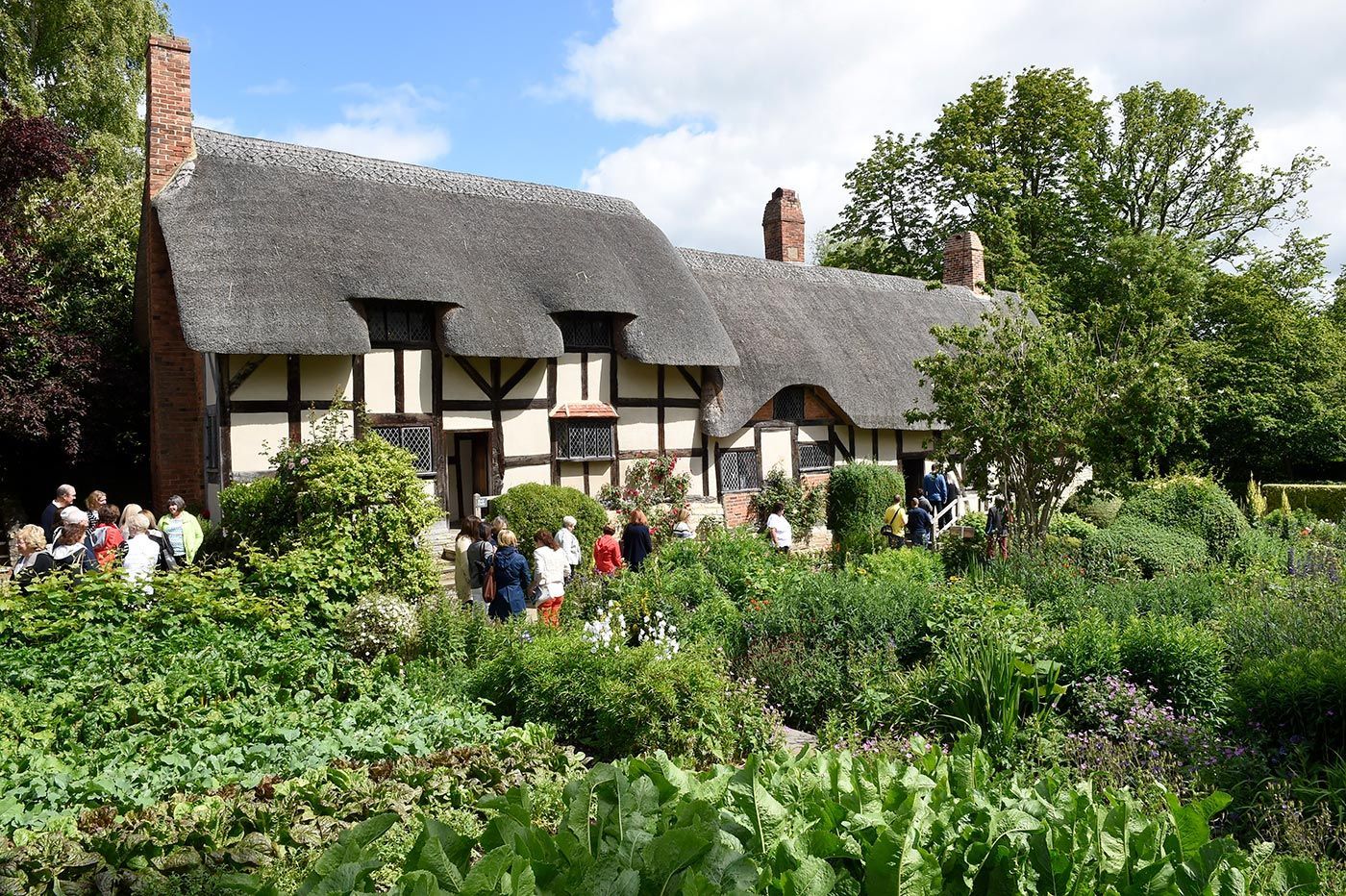 Thatched-roof cottage with garden and people gathered in front. Sunny day.