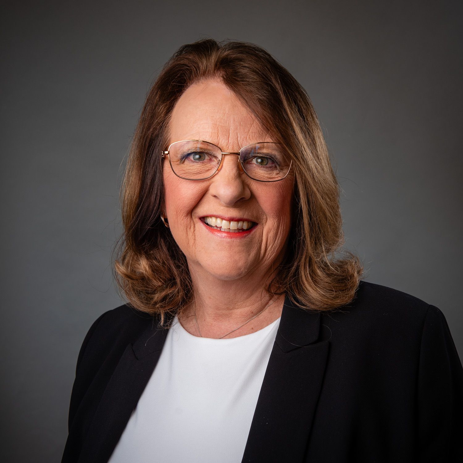 Woman with glasses smiles, wearing a black blazer and white top, against a gray background.