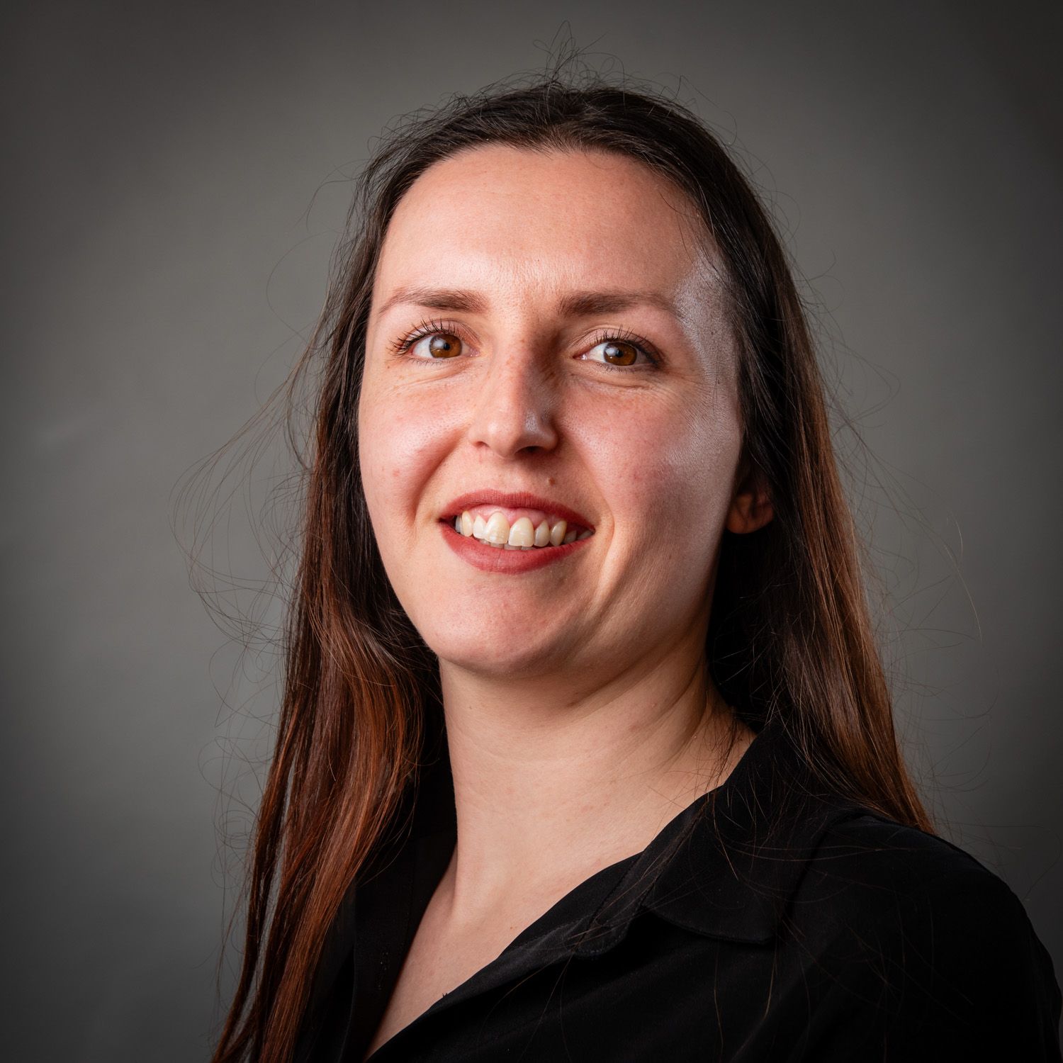Woman with brown hair smiles, wearing a black shirt, against a gray backdrop.
