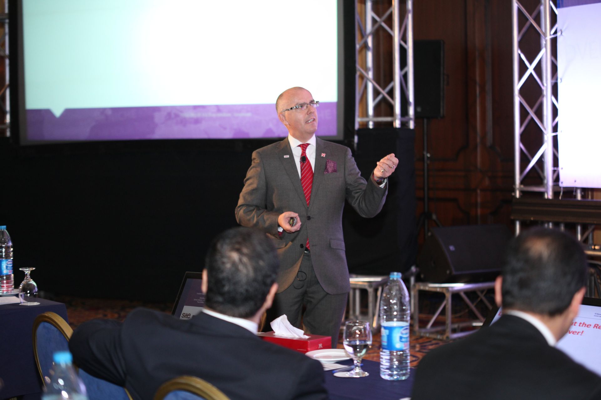 Man in suit giving presentation at a conference, gesturing towards a screen; audience seated at tables.