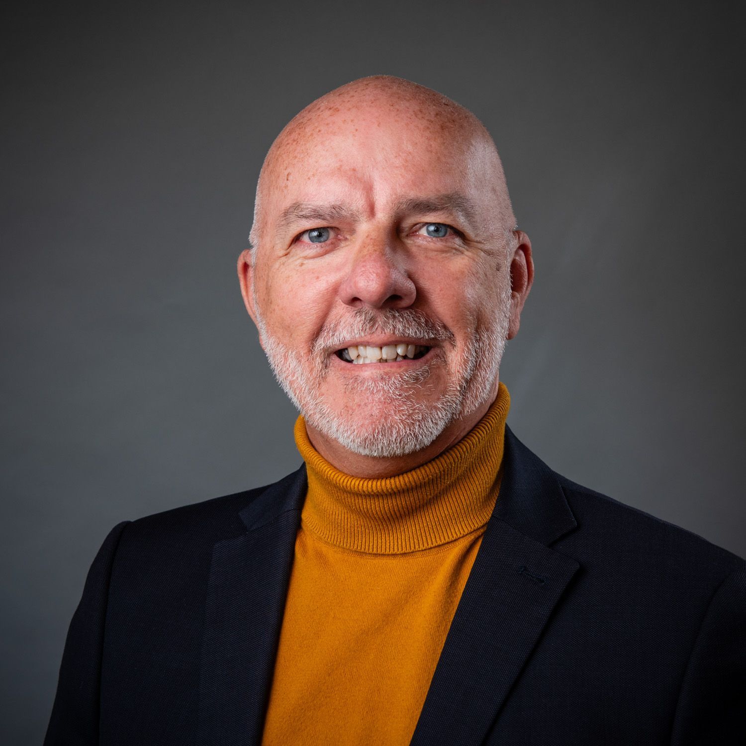 Bald man smiles, wearing orange turtleneck and dark blazer against gray background.