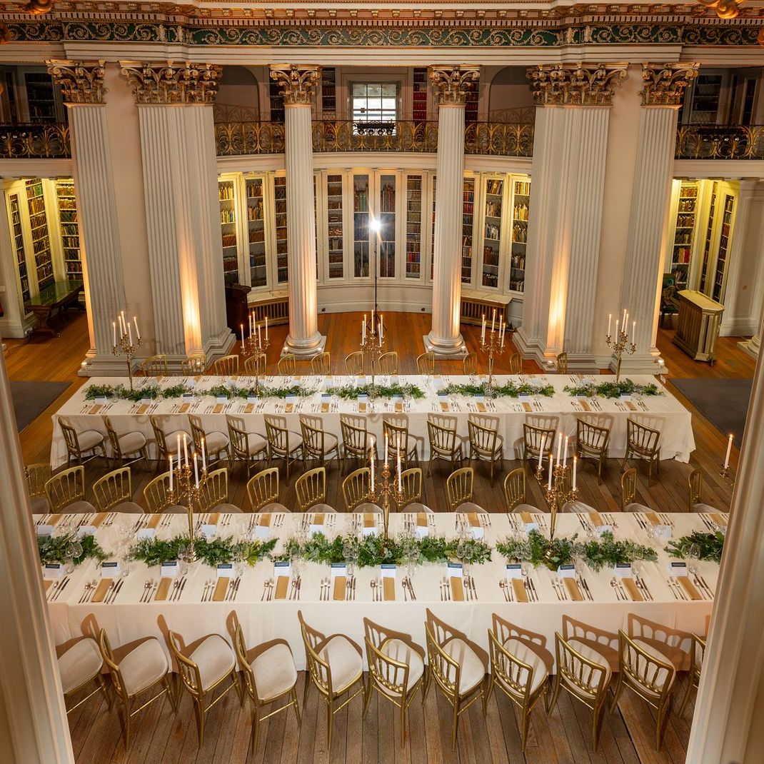 Formal dining hall set for event, long tables, gold chairs, lit by candles, columns in the background.