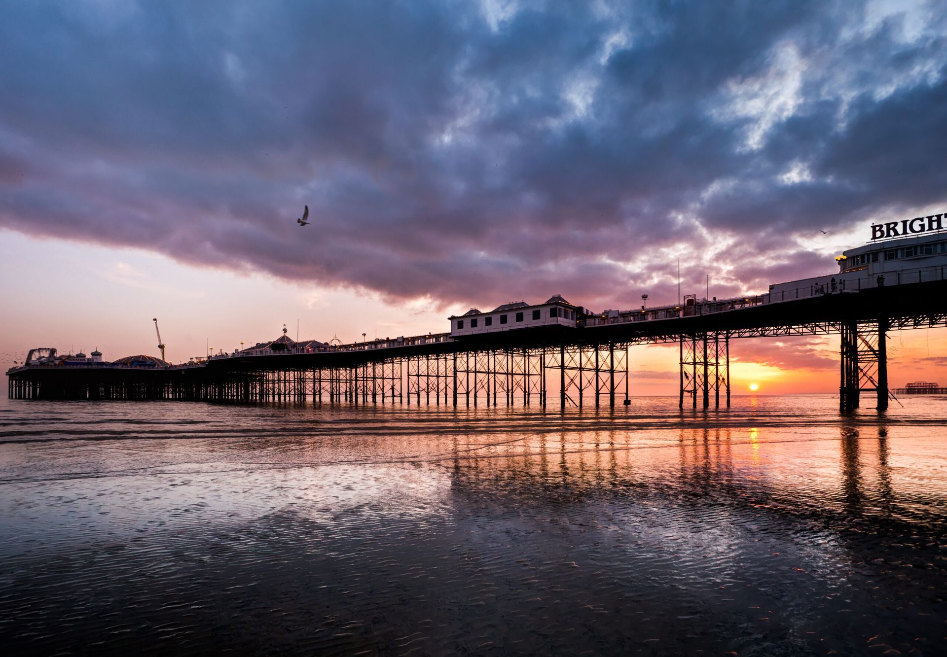 Brighton Pier at sunset, silhouetted against a colorful sky. Reflections in the wet sand.
