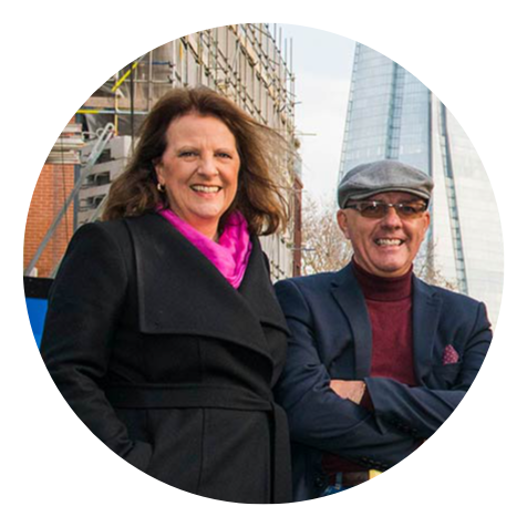 Woman in black coat and man in suit stand in front of buildings, smiling.