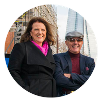Woman in black coat and man in suit stand in front of buildings, smiling.