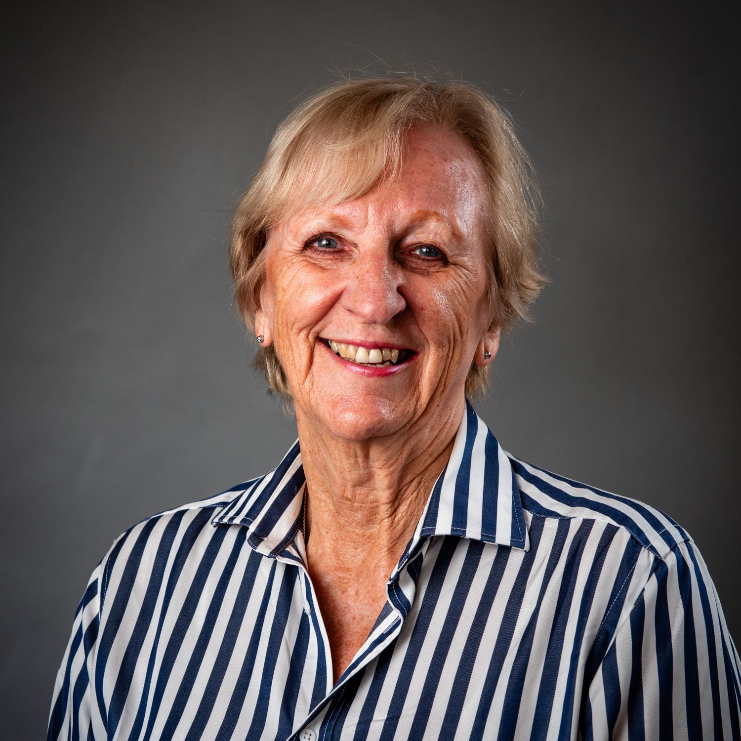 Woman with short blonde hair smiles, wearing a striped blue and white shirt, against a gray background.