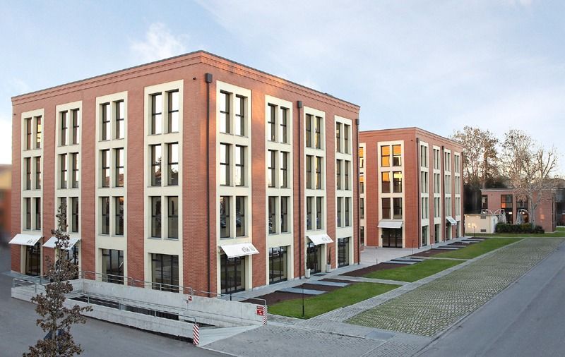 Edifici per uffici in mattoni con numerose finestre, prato verde e un marciapiede pavimentato.