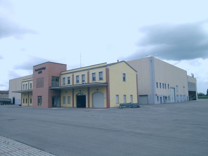 Un grande edificio industriale, color beige e salmone, con uno sfondo di cielo nuvoloso.