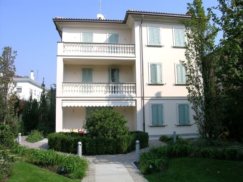 Edificio beige a tre piani con balconi, persiane azzurre e giardino ben curato.