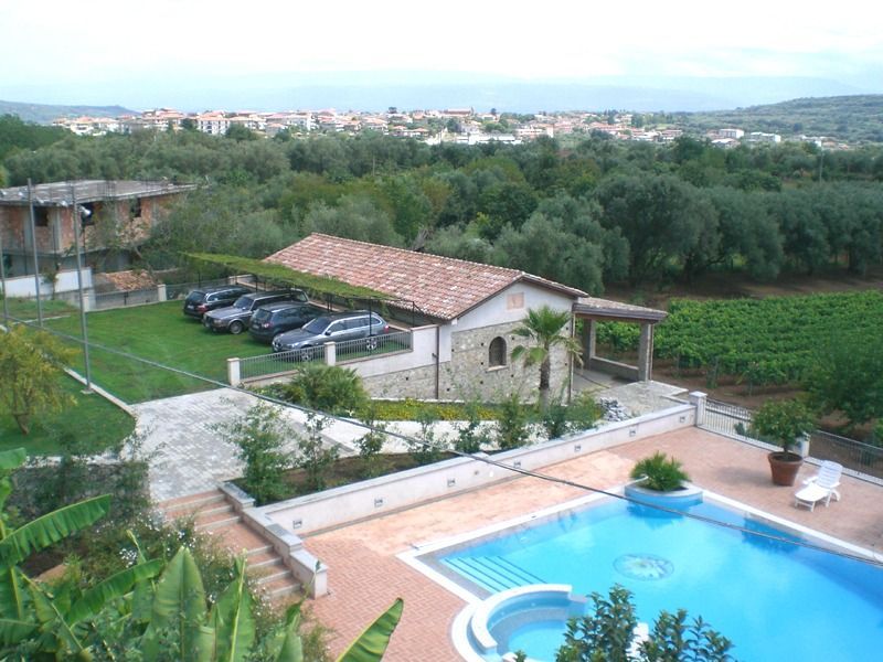 Piscina in un ambiente rurale con edifici, automobili, alberi e una città lontana sotto un cielo nuvoloso.