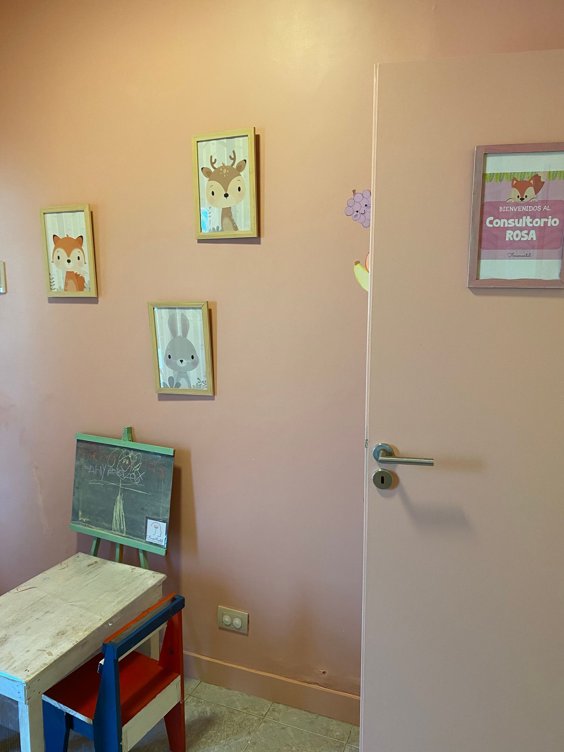 Habitación rosa con arte animal enmarcado, mesa pequeña, silla y una puerta cerrada.